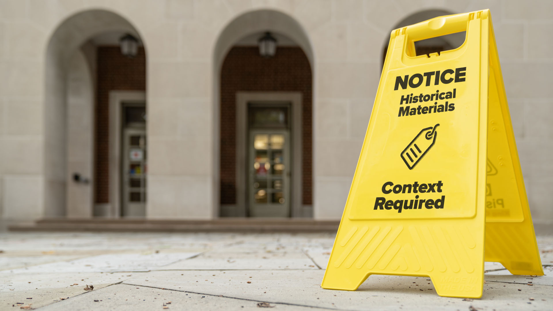 A photo illustration of a small easel-style sign typically used to warn of wet floors in front of King Library. The sign reads: Notice. Historical Materials. Context Required. The sign also has an icon of a tag on it.