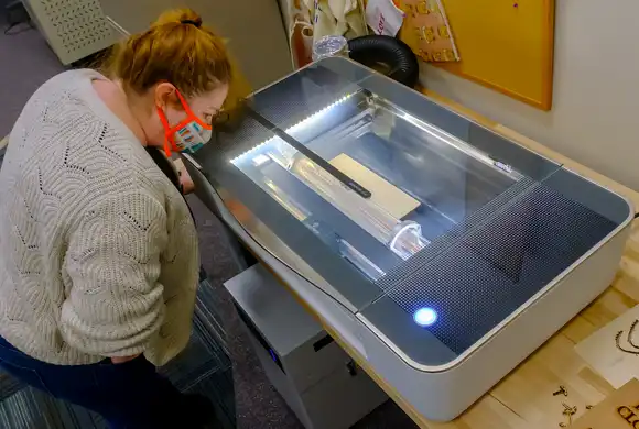 A woman wearing a mask looks down at a Glowforge laser cutter and engraver as it operates.