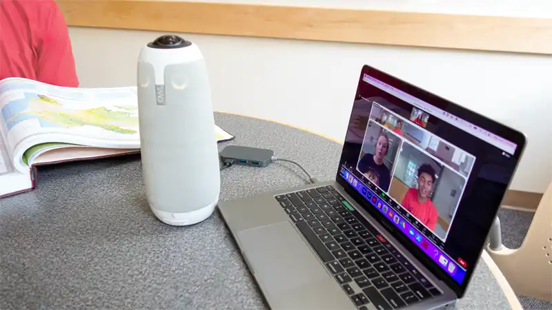 A stuffed animal toy owl sits next to a Meeting Owl device in front of a laptop depicting a Zoom meeting between students in different study rooms.