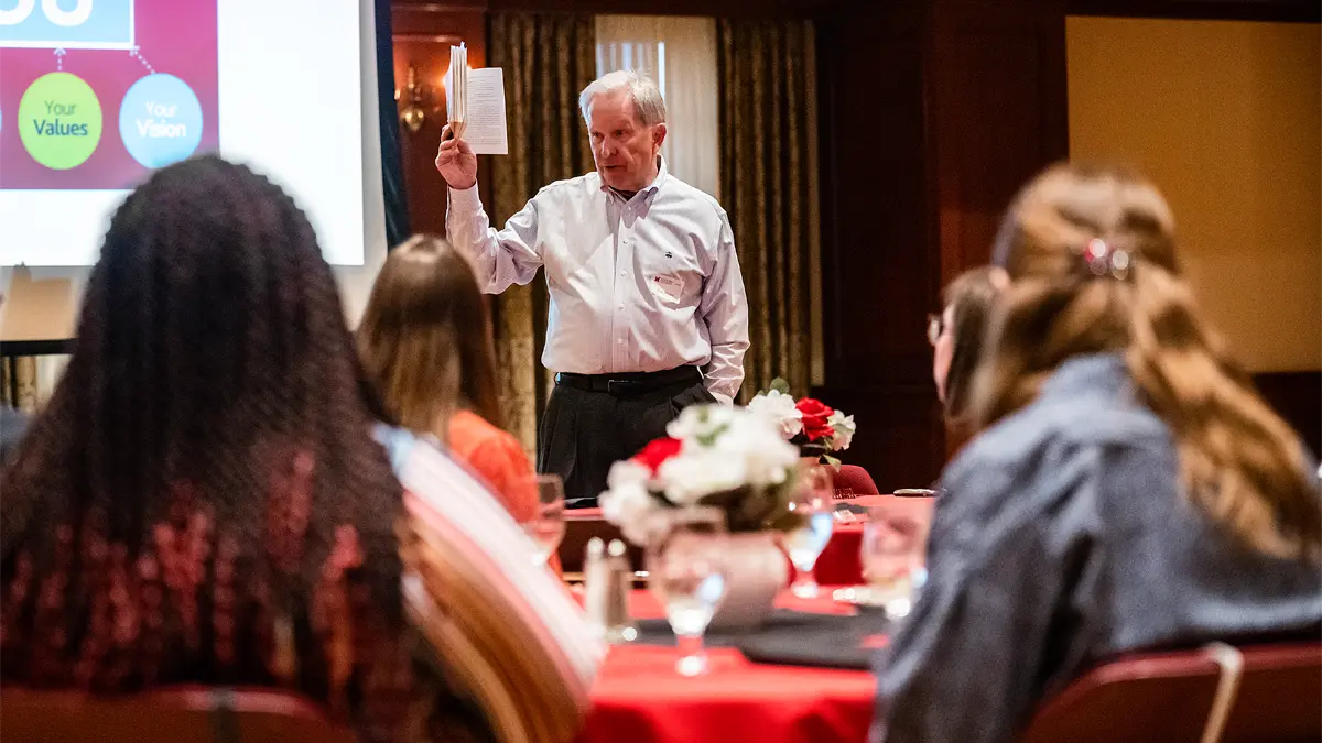 Tom Heuer holds up a book as he speaks to students seated at tables in the Shriver Center at Miami University. A projector screen behind him displays a slide with the words: Your Values and Your Vision.