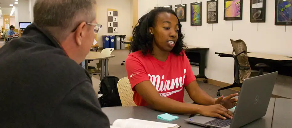 A young woman works on a laptop while a librarian looks on